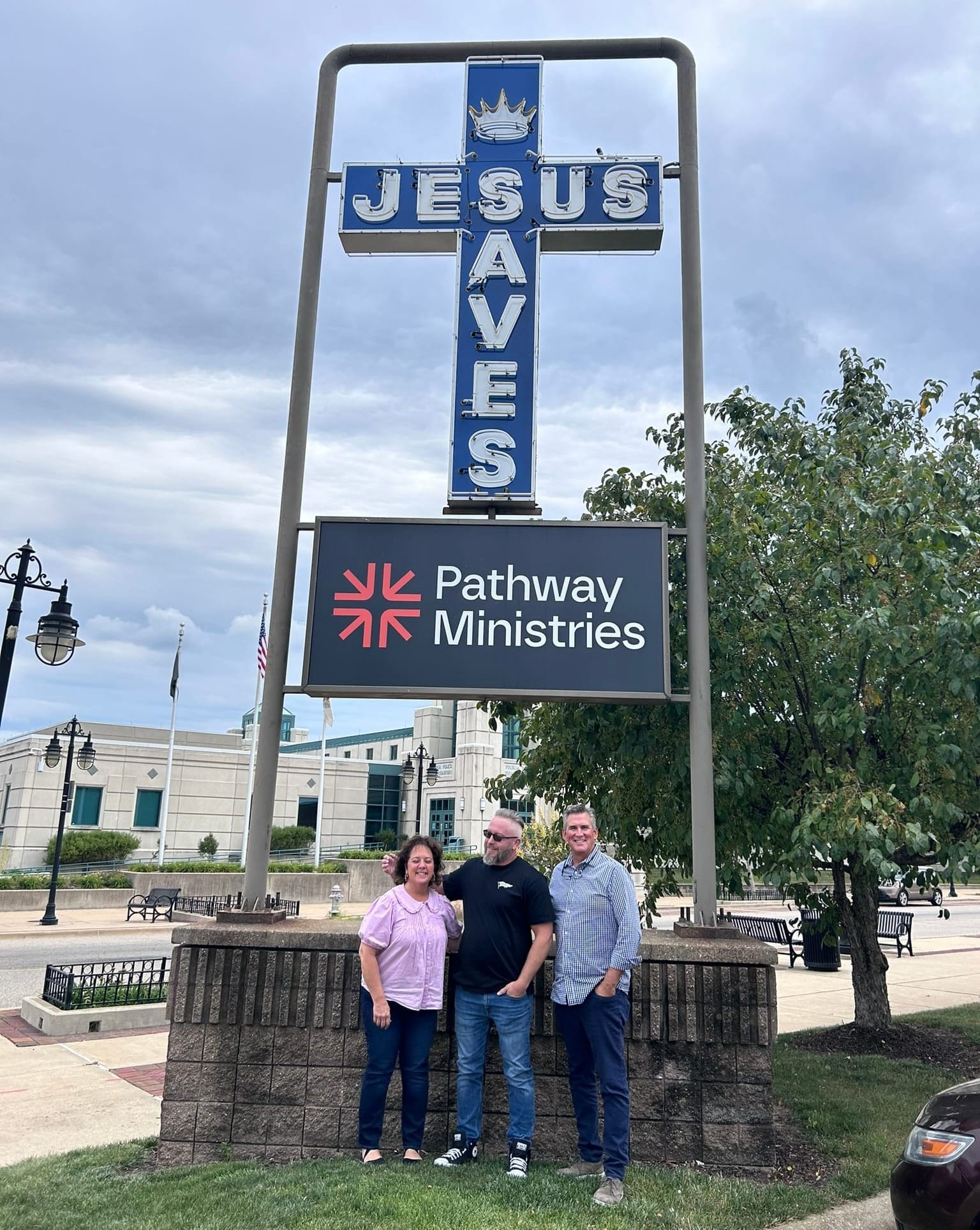 Three people stand in front of a sign for Pathway Ministries and a cross that says Jesus Saves.